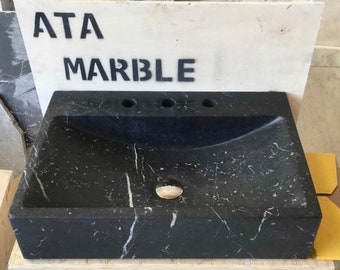 May include: Black marble bathroom sink with three holes for faucets. The sink is rectangular and has a curved interior. The sink is made by ATA Marble.