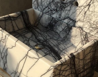 May include: A rectangular marble sink with a backsplash. The sink is white with dark gray veining, and has a rectangular basin. The backsplash has three circular faucet holes. The sink is made of stone and is likely for a bathroom.