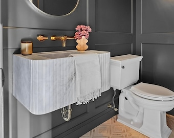May include: A white marble sink with a gold faucet and a white towel hanging on the side. The sink is in a bathroom with dark gray walls and a white toilet.