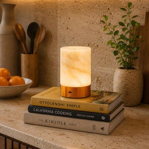 May include: A cylindrical lamp with a glowing, marbled white stone shade and a metallic copper base, resting on a stack of three books titled Ottolenghi Simple, California Cooking by Sheilah Lukins, and The Kinfolk Table, emitting a warm light.
