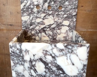 May include: A white marble sink with a black and purple marbling pattern. The sink has a square shape and a backsplash with three holes.