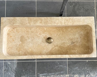 May include: A rectangular stone sink with a round drain in the center. The sink is made of a light beige stone with a marbled pattern.