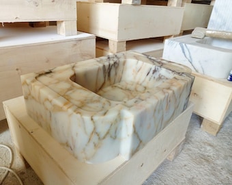 Puede incluir: Un lavabo grande de mármol blanco con vetas marrones, sentado en una caja de madera. El lavabo es rectangular con un borde delantero redondeado y una abertura cuadrada en el centro.
