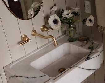 May include: A wall-mounted white marble sink with prominent black veining, featuring a brass faucet and two matching brass handles. A glass vase with white anemone flowers sits on the sink ledge next to a small bowl, below an oval mirror.