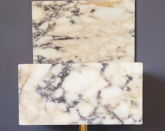 May include: A white marble wall shelf with a rectangular shape and a gold metal bracket. The shelf has a unique design with two layers, creating a tiered effect.