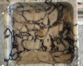 May include: A white marble sink with a brown and gray veining pattern. The sink has a round drain hole in the center.