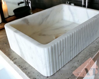 May include: A rectangular white marble sink with vertical grooved sides. The sink has a smooth interior and sits on a speckled countertop. The bathroom fixture is a modern design.
