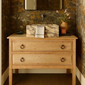 Könnte beinhalten: Ein helles Holzwaschbeckenunterschrank mit zwei Schubladen und Ringgriffen. Ein Marmorwaschbecken steht oben, mit einer kleinen Vase mit Blumen und einer Seifenschale. Der Hintergrund zeigt eine Tapete mit Blättermuster.