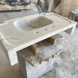 Puede incluir: Un lavabo de ba&ntilde;o de m&aacute;rmol blanco con una sola cuenca y tres agujeros para accesorios. La encimera se est&aacute; construyendo sobre un marco de madera.