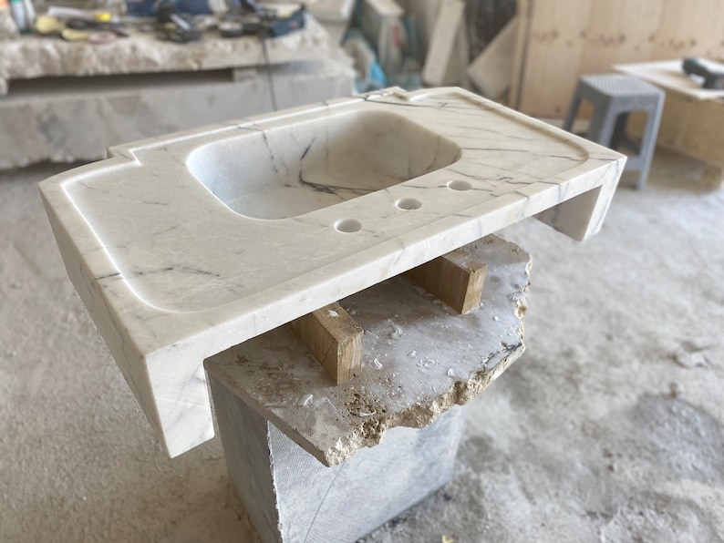 Puede incluir: Un lavabo de ba&ntilde;o de m&aacute;rmol blanco con una sola cuenca y tres agujeros para accesorios. La encimera se est&aacute; construyendo sobre un marco de madera.