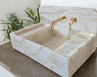 Puede incluir: Un lavabo de baño de mármol blanco con un grifo dorado y dos pomos dorados. El lavabo tiene forma rectangular y un borde elevado.