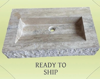 May include: A rectangular stone sink with a central drain hole. The sink has a recessed basin and a rough, textured exterior. The color is a mix of beige and gray. The text "READY TO SHIP" is below the sink.