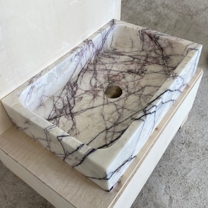 May include: A white marble sink with purple veins. The sink is rectangular and has a single drain hole. The sink is mounted on a wooden base.