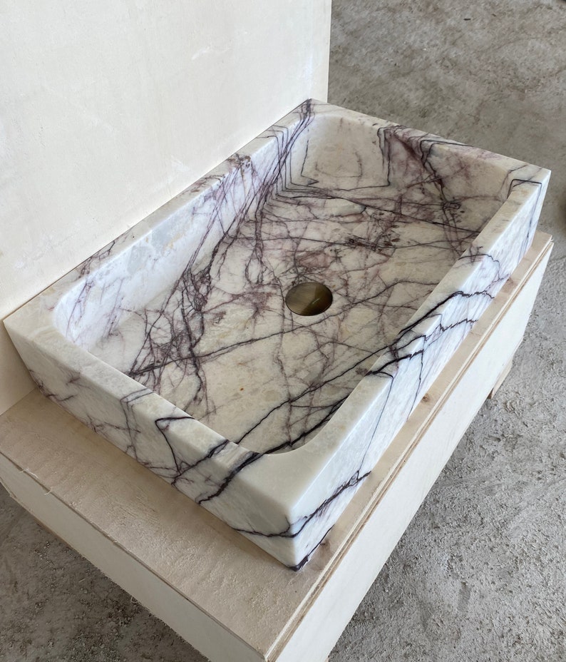 May include: A white marble sink with purple veins. The sink is rectangular and has a single drain hole. The sink is mounted on a wooden base.