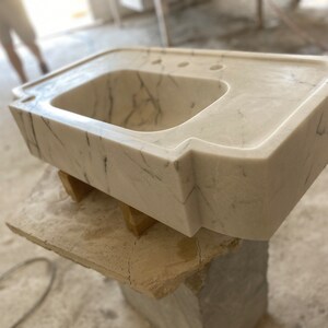 Puede incluir: Un lavabo de ba&ntilde;o de m&aacute;rmol blanco con un dise&ntilde;o moderno y &uacute;nico. El lavabo tiene una forma rectangular con un borde frontal curvo y una sola cuenca. El lavabo est&aacute; sentado sobre una base de madera.