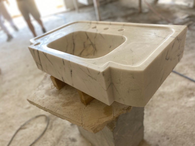 Puede incluir: Un lavabo de ba&ntilde;o de m&aacute;rmol blanco con un dise&ntilde;o moderno y &uacute;nico. El lavabo tiene una forma rectangular con un borde frontal curvo y una sola cuenca. El lavabo est&aacute; sentado sobre una base de madera.