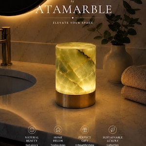 May include: A cylindrical table lamp featuring a translucent green onyx stone body with natural veining, illuminated from within, and set upon a solid metallic gold base. The lamp is positioned on a marble countertop with text reading ATAMARBLE.