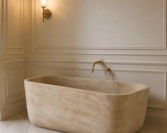 May include: A rectangular, beige stone bathtub with rounded edges is the focal point of this bathroom. A gold faucet and wall sconce add a touch of luxury. The walls are a neutral color with decorative molding, creating a spa-like atmosphere.