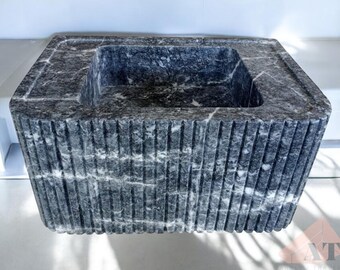 Può includere: Un lavandino in pietra di colore grigio scuro con un bacino rettangolare e un esterno scanalato. Il lavandino è realizzato in pietra naturale e ha un aspetto rustico e artigianale.