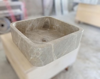 May include: A square, stone bathroom sink with a light gray marble pattern. The sink has a rounded rim and a drain hole in the center. The sink is sitting on a white surface.