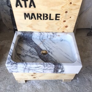 May include: A white marble sink with gray veining, set on a wooden base with the text "ATA MARBLE" on the side.