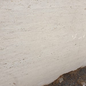 May include: A close-up of a large, light beige travertine slab with a smooth, polished surface. The slab has a natural, veined pattern and a slightly textured finish.