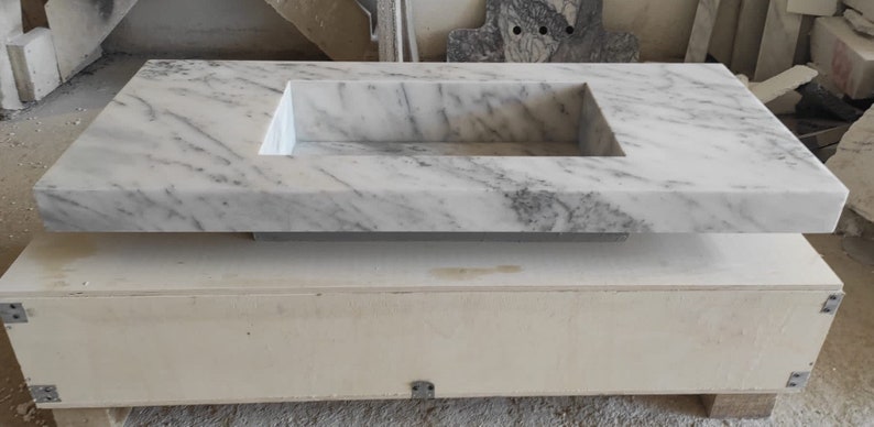 Puede incluir: Un lavabo de ba&ntilde;o de m&aacute;rmol blanco con un recorte rectangular para la cuenca del lavabo. La encimera est&aacute; sentada sobre una base de madera.