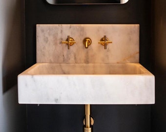 May include: A white marble bathroom sink with a gold faucet and drain. The sink is mounted on a black wall with a gold mirror above it.