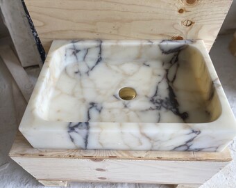 May include: A white marble sink with grey veining, set in a wooden crate. The sink has a round, brass drain.
