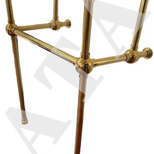 Könnte beinhalten: Goldfarbener Waschbeckenständer aus Metall mit drei Beinen und einer horizontalen Stange. Der Ständer ist aus poliertem Metall gefertigt und hat ein traditionelles Design.