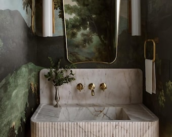 May include: A white marble bathroom sink with a gold faucet and a gold-framed mirror reflecting a landscape painting. The sink is set against a dark green wall with a green and blue landscape wallpaper.