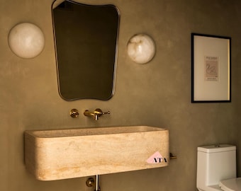 May include: A modern bathroom with a beige stone sink, gold fixtures, and a matching mirror. Two round wall sconces flank the mirror. A framed Picasso print and a white toilet complete the space. The walls are painted in a muted olive green.