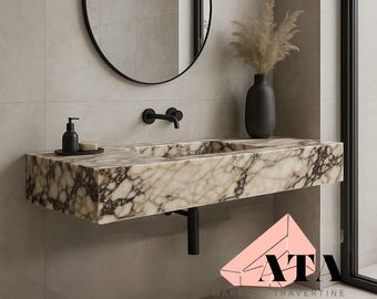 Puede incluir: Un lavabo de baño de mármol con vetas marrones y beige. El lavabo rectangular está montado en la pared con fontanería negra. Un grifo negro, espejo y dispensador de jabón completan el diseño minimalista. También hay un jarrón con plantas secas.