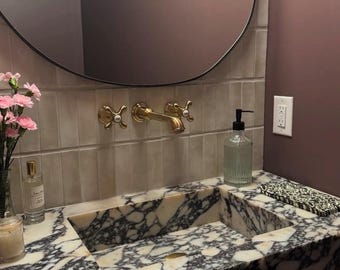 May include: A white marble bathroom vanity with a black veined pattern. The vanity has a rectangular sink and a gold faucet with two knobs. A clear glass soap dispenser is on the counter next to the sink.