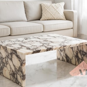 May include: A rectangular coffee table with a marble-like pattern in shades of cream and brown. The table is supported by thick, solid sides, and is placed in front of a cream-colored sofa. The table's dimensions are approximately 48 inches long, 24 inches wide, and 16 inches high.