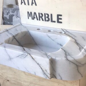 Puede incluir: Un lavabo de ba&ntilde;o de m&aacute;rmol blanco con un borde frontal curvo y tres agujeros para grifos. El lavabo est&aacute; sentado sobre una base de madera.