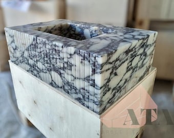 Puede incluir: Una pila de azulejos de mármol blanco con vetas grises, dispuestos en forma rectangular, con un recorte en el centro. Los azulejos están apilados en una caja de madera.