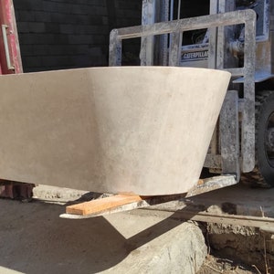 May include: A large, light-colored bathtub being loaded onto a forklift. The bathtub is made of a smooth, textured material and has a rounded shape. The forklift is yellow and has a black cage.