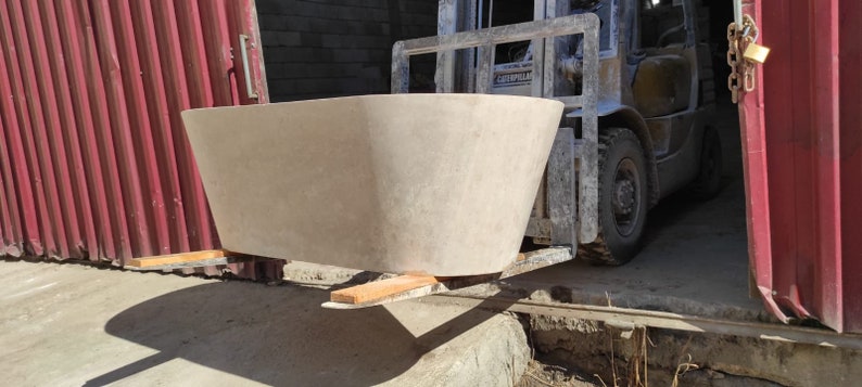 May include: A large, light-colored bathtub being loaded onto a forklift. The bathtub is made of a smooth, textured material and has a rounded shape. The forklift is yellow and has a black cage.