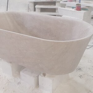 May include: A large, oval-shaped bathtub made of light-colored stone. The bathtub is sitting on blocks of stone and is in a workshop setting.