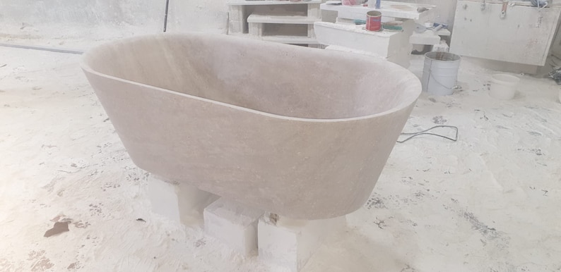 May include: A large, oval-shaped bathtub made of light-colored stone. The bathtub is sitting on blocks of stone and is in a workshop setting.