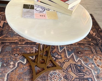 May include: A round, white marble-topped coffee table with a gold-colored metal base. The table holds a stack of books, a teacup and saucer with blue floral designs, and a small card with sample images. The table sits on a patterned rug.