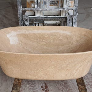 May include: A large, oval-shaped bathtub made of light brown travertine stone. The bathtub is sitting on a concrete floor.