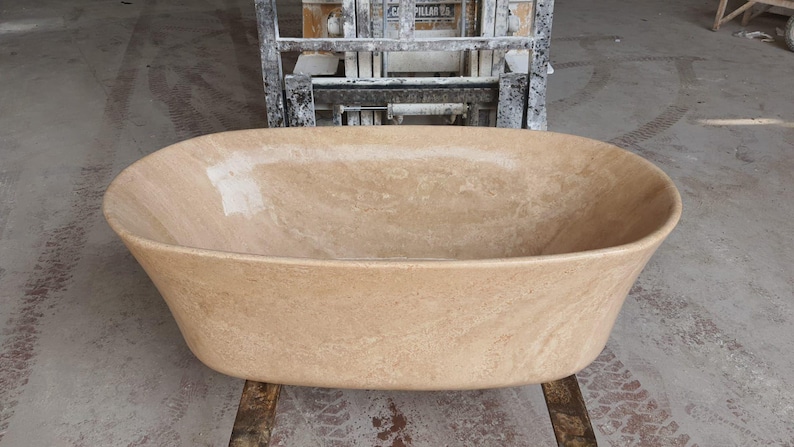 May include: A large, oval-shaped bathtub made of light brown travertine stone. The bathtub is sitting on a concrete floor.