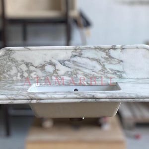May include: A marble countertop with a built-in rectangular sink. The countertop has a white and gray veined pattern, with a backsplash. The text "ATAM MARBLE" is visible on the countertop.