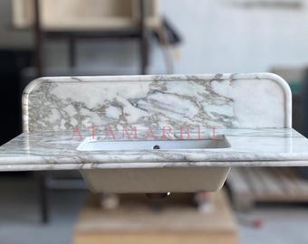 May include: A marble countertop with a built-in rectangular sink. The countertop has a white and gray veined pattern, with a backsplash. The text "ATAM MARBLE" is visible on the countertop.