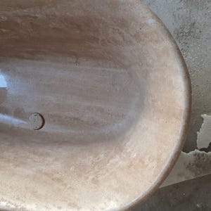 May include: A close-up of a light brown oval bathtub with a drain in the bottom. The bathtub is made of a natural stone material with a smooth, polished finish.