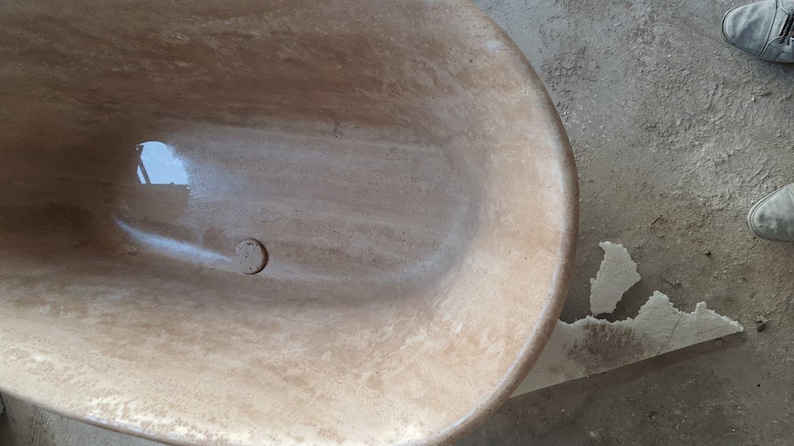 May include: A close-up of a light brown oval bathtub with a drain in the bottom. The bathtub is made of a natural stone material with a smooth, polished finish.