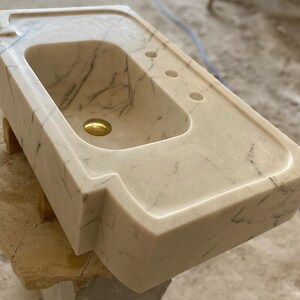 Puede incluir: Un lavabo de ba&ntilde;o de m&aacute;rmol blanco con un dise&ntilde;o moderno y &uacute;nico. El lavabo tiene una forma rectangular con bordes redondeados y una sola cuenca grande. El lavabo est&aacute; hecho de m&aacute;rmol de alta calidad y tiene un acabado liso y pulido.