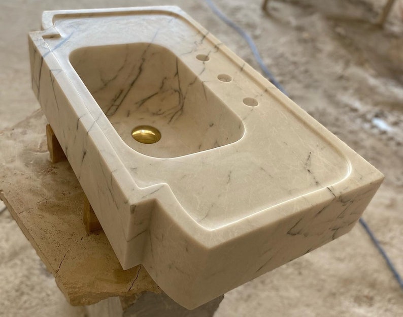 Puede incluir: Un lavabo de ba&ntilde;o de m&aacute;rmol blanco con un dise&ntilde;o moderno y &uacute;nico. El lavabo tiene una forma rectangular con bordes redondeados y una sola cuenca grande. El lavabo est&aacute; hecho de m&aacute;rmol de alta calidad y tiene un acabado liso y pulido.
